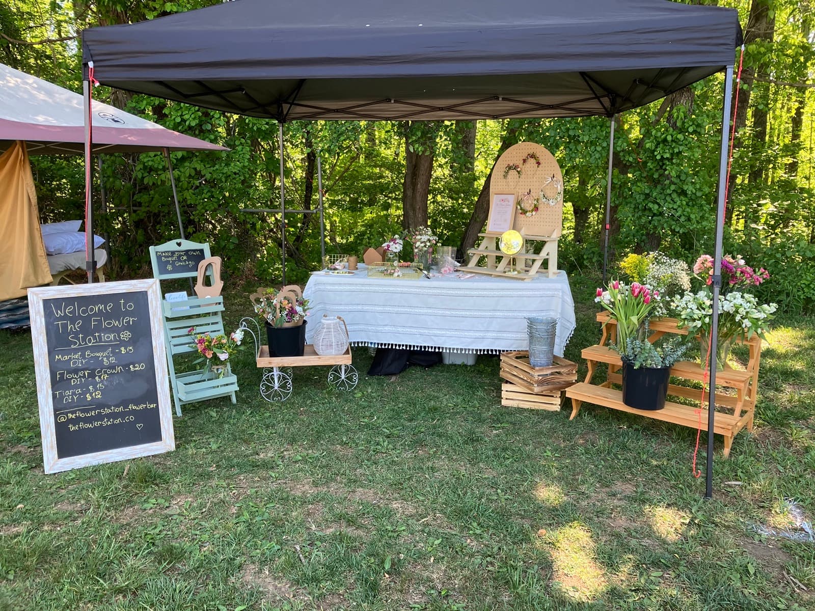 The Flower Station full booth setup at an outdoor Asheville event — welcome chalkboard sign and wooden display shelves