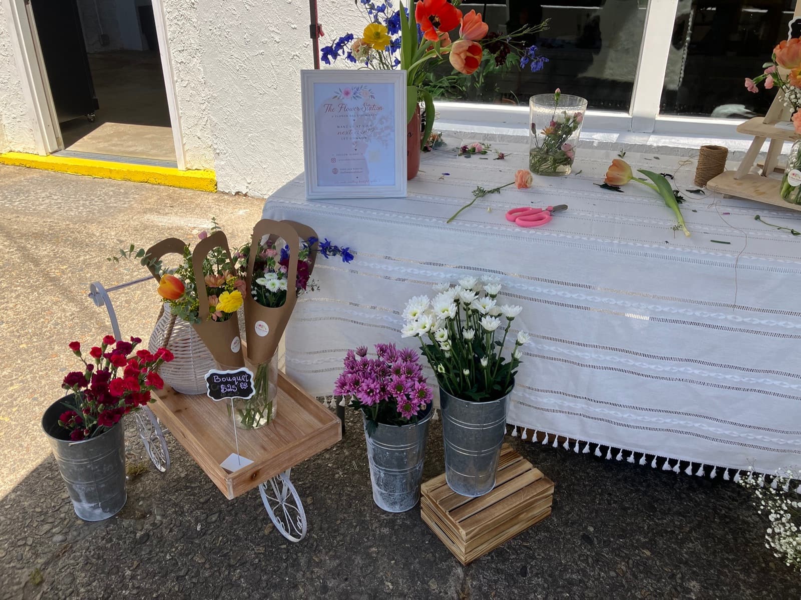 Outdoor event setup with white bicycle cart, fresh flower buckets, and styled table